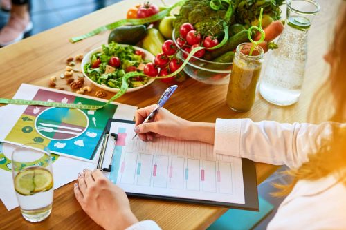 Woman dietitian in medical uniform with tape measure working on a diet plan sitting with different healthy food ingredients in the green office on background. Weight loss and right nutrition concept