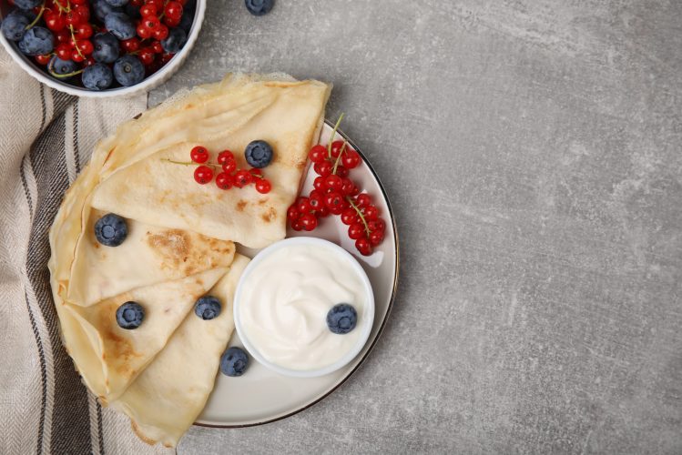 Delicious crepes with natural yogurt, blueberries and red currants on grey table, flat lay. Space for text